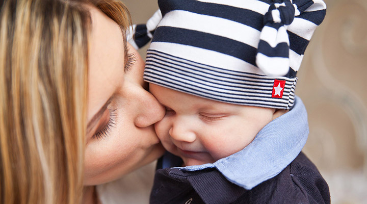 Baby mit Mütze bekommt einen Kuss von Mama. Die richtige Mützengröße für Ihr Baby hat Sie vorher durch Messen des Kopfumfangs und einen Blick in die Baby Mützengrößen Tabelle herausgefunden.